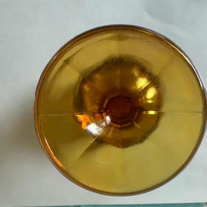 May include: Overhead view of a golden-colored glass bowl. The bowl has a wide, shallow shape with a rounded base and a flared rim. The glass has a reflective, shiny surface, and the color is a warm, amber hue.