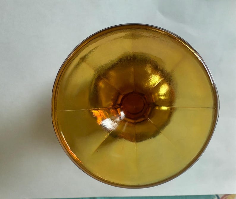 May include: Overhead view of a golden-colored glass bowl. The bowl has a wide, shallow shape with a rounded base and a flared rim. The glass has a reflective, shiny surface, and the color is a warm, amber hue.