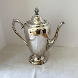 以下が含まれることがあります： 装飾的な花柄のデザインが施された銀色のコーヒーポット。ポットは、湾曲した注ぎ口、ハンドル、装飾的なつまみ付きの蓋を備えています。台座の上に置かれ、白い表面に展示されています。