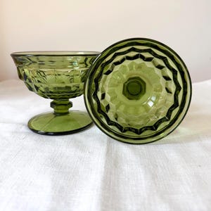 May include: Two green glass dessert dishes. One is upright, the other is upside down. The dishes have a textured, geometric pattern on the bowl and a pedestal base. The glass has a slight olive green hue.