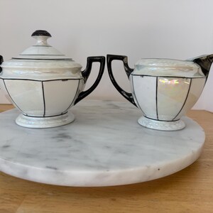 Puede incluir: Un azucarero vintage con tapa y un juego de crema. Las piezas de porcelana blanca tienen detalles y asas negros. El juego se exhibe en una bandeja redonda de m&aacute;rmol. El azucarero tiene un pomo negro en la tapa.