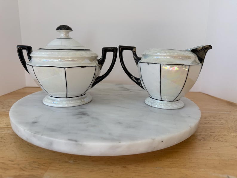 Puede incluir: Un azucarero vintage con tapa y un juego de crema. Las piezas de porcelana blanca tienen detalles y asas negros. El juego se exhibe en una bandeja redonda de m&aacute;rmol. El azucarero tiene un pomo negro en la tapa.