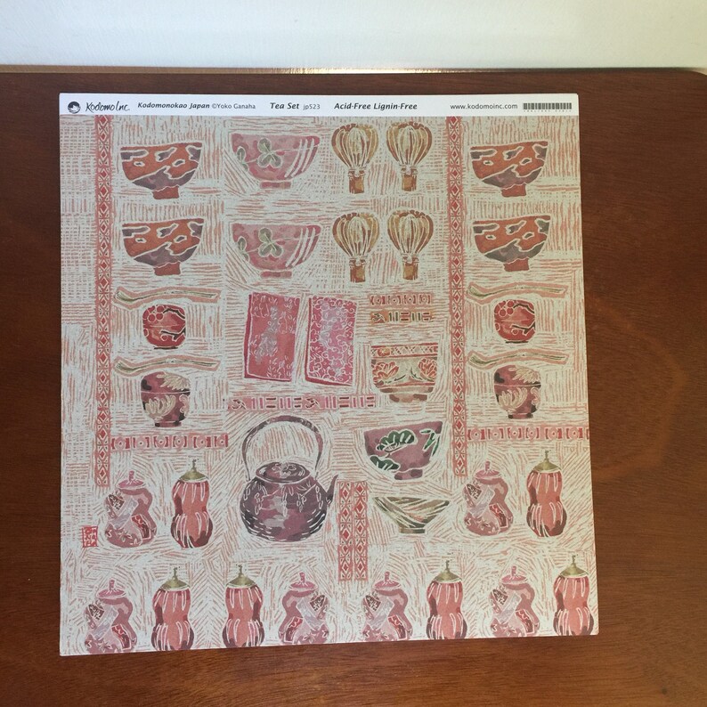 Puede incluir: Una hoja de papel decorativo con un tema de juego de t&eacute; japon&eacute;s. El papel presenta un patr&oacute;n repetitivo de tazas de t&eacute;, teteras y otros art&iacute;culos relacionados con el t&eacute; en tonos de rojo, marr&oacute;n y blanco. El texto "Tea Set 523 Acid-Free Lignin-Free" est&aacute; impreso en la parte superior de la hoja.
