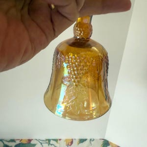 May include: An amber-colored glass bell with a decorative grape design. The bell is held by a hand, showcasing its iridescent finish. The bell has a rounded base and a long stem.