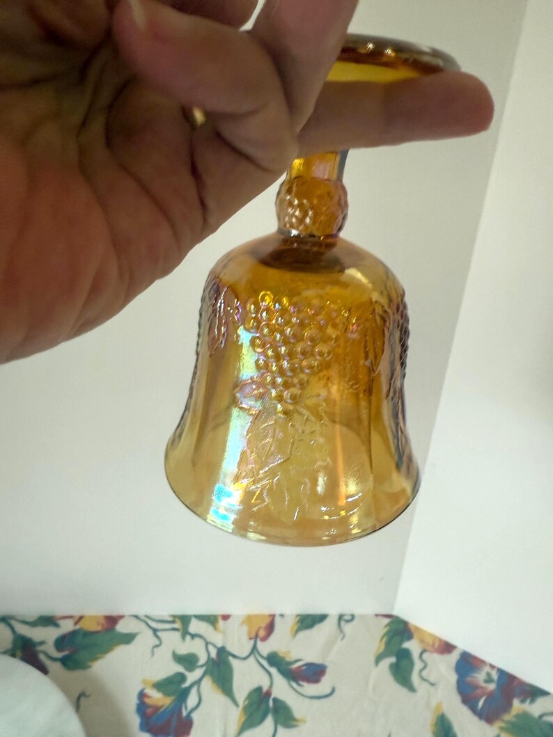 May include: An amber-colored glass bell with a decorative grape design. The bell is held by a hand, showcasing its iridescent finish. The bell has a rounded base and a long stem.
