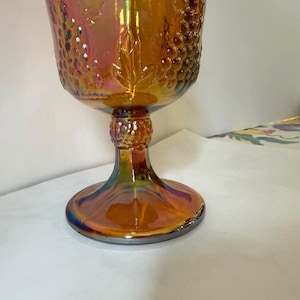 May include: An iridescent orange goblet with a textured grape design on the bowl. The stem and base have a similar iridescent finish, with a round base. The glass reflects a range of colors, including gold, purple, and blue.