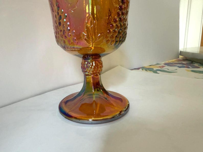 May include: An iridescent orange goblet with a textured grape design on the bowl. The stem and base have a similar iridescent finish, with a round base. The glass reflects a range of colors, including gold, purple, and blue.