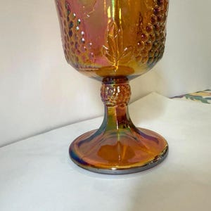May include: An iridescent orange glass goblet with a detailed grape and leaf design. The stem and base have a similar iridescent finish, creating a vintage aesthetic. The goblet is likely used for serving beverages.