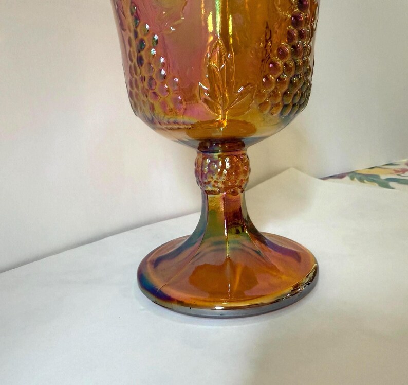 May include: An iridescent orange glass goblet with a detailed grape and leaf design. The stem and base have a similar iridescent finish, creating a vintage aesthetic. The goblet is likely used for serving beverages.