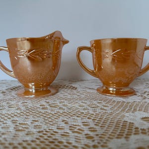 May include: Two iridescent orange glass serving pieces. One is a creamer with a spout and handle, and the other is a sugar bowl with two handles. Both pieces have a leaf design and sit on a pedestal base.