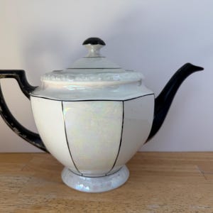 Puede incluir: Una tetera de porcelana blanca con asa, boquilla y detalles en negro. La tetera tiene una tapa con un pomo negro y est&aacute; decorada con l&iacute;neas negras. El acabado nacarado le da un aspecto vintage. Un art&iacute;culo decorativo para la cocina.
