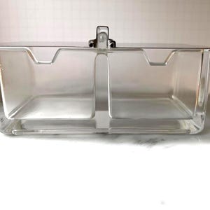 Pode incluir: Um recipiente retangular de vidro transparente com compartimento duplo, uma divisória central e uma tampa metálica plana e refletora. A tampa inclui uma pequena pega vertical centralizada para levantar, com duas secções abertas.