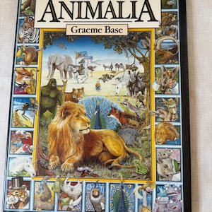May include: A hardcover book titled "Animalia" by Graeme Base. The cover features a vibrant illustration of a lion, peacock, and various other animals. The book is framed with a black border and includes the title in large, bold letters.