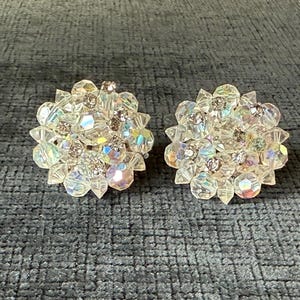 Puede incluir: Un par de pendientes de clip de cristal transparente y facetado. Cada pendiente es redondo, con un grupo central de cristales m&aacute;s peque&ntilde;os rodeado de cristales m&aacute;s grandes y puntiagudos. Los cristales tienen una cualidad iridiscente.