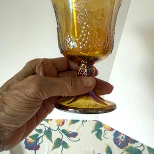 May include: An iridescent amber-colored goblet with a grape vine design. The glass has a textured surface and a decorative stem. The goblet is held in a hand, showcasing its details and the play of light on its surface.