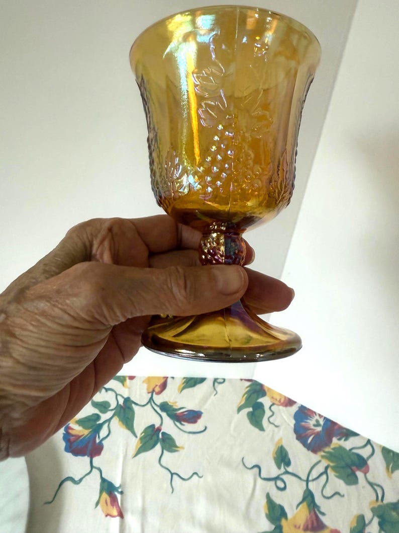 May include: An iridescent amber-colored goblet with a grape vine design. The glass has a textured surface and a decorative stem. The goblet is held in a hand, showcasing its details and the play of light on its surface.