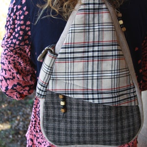 May include: A plaid fabric sling bag with a beige, black, and red pattern. The bag has a triangular shape with a flap and wooden button details. The bottom section is a darker gray plaid. The bag is worn over the shoulder.