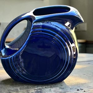 May include: A deep cobalt blue ceramic pitcher featuring a rounded, disc-like body with concentric circular embossed rings. The vessel includes a curved handle integrated into the side and a prominent pouring spout at the top rim.