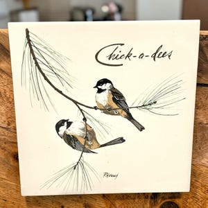 May include: A square white ceramic tile featuring a hand-painted illustration of two black-capped chickadees perched on a pine branch. The text Chick-a-dees is written in black script in the upper right corner, with the artist signature P. Horned below.