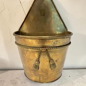 May include: A wall-mounted brass planter featuring a semi-circular bucket shape with a flat back panel. The exterior is decorated with a horizontal twisted rope trim that includes two hanging tassels centred on the front of the brass vessel.