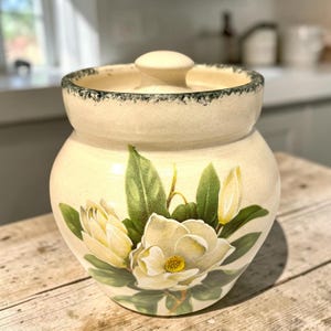 May include: A cream-colored ceramic canister with a matching lid, featuring a hand-painted design of white magnolia flowers and green leaves. The rim of the lid and the top edge of the canister have a dark, speckled, distressed finish.