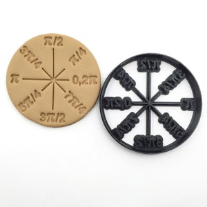 Könnte beinhalten: Ein hellbrauner, kreisförmiger Keks mit einem Kompass-Design, das mathematische Symbole und Brüche enthält. Daneben befindet sich ein schwarzer Kunststoff-Ausstecher mit demselben Design. Der Ausstecher wird verwendet, um das Design auf den Keks zu prägen.