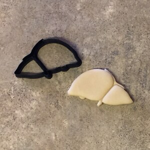 May include: A black cookie cutter in the shape of a liver sits next to a cookie in the same shape. The cookie is a light beige color. The background is a mottled gray.