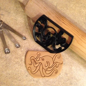 Könnte beinhalten: Ein schwarzer Ausstecher in Form eines Oktopus und ein Keks mit einem Oktopus-Design. Ein Nudelholz aus Holz und Messlöffel sind ebenfalls im Bild. Der Ausstecher ist aus Kunststoff.