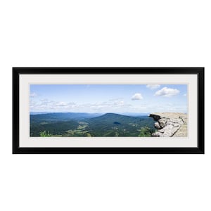 May include: A panoramic view from a clifftop overlooking a vast, green valley. The sky is blue with fluffy white clouds. The cliff edge is rocky and appears to be jutting out over the valley.