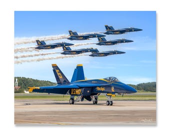 May include: A blue and yellow F/A-18 Super Hornet jet aircraft, part of the U.S. Navy Blue Angels flight demonstration squadron, is on the runway with five other jets flying in formation overhead.