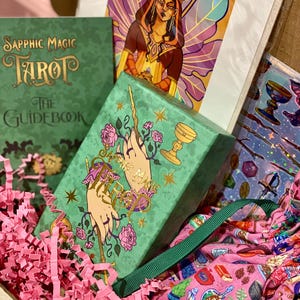 May include: A gift box containing a green tarot card box with gold lettering and illustrations, a green guidebook, and a colorful illustration of a woman with wings. The box is filled with pink paper filler and a pink patterned fabric.