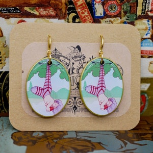 Peut inclure: Une paire de boucles d'oreilles en or avec des pendentifs de forme ovale représentant une illustration colorée d'une femme suspendue la tête en bas à une branche d'arbre. La femme porte des bas rayés rose et blanc et un haut rayé rouge et blanc. L'arrière-plan est un paysage vert et bleu.