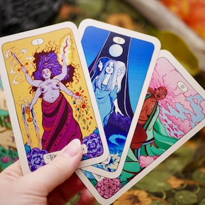 May include: Three tarot cards featuring colorful illustrations are held in a hand. The cards depict figures with vibrant colors, including a woman with multiple arms, a blue-toned scene with three figures, and a figure in green and pink.