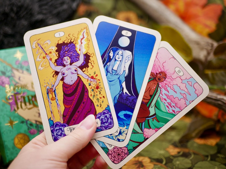May include: Three tarot cards featuring colorful illustrations are held in a hand. The cards depict figures with vibrant colors, including a woman with multiple arms, a blue-toned scene with three figures, and a figure in green and pink.