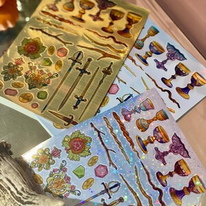 May include: Sheets of decorative stickers featuring tarot card suit symbols. The stickers include chalices, swords, wands, and pentacles in gold, blue, and iridescent finishes. The stickers are arranged on a light background.