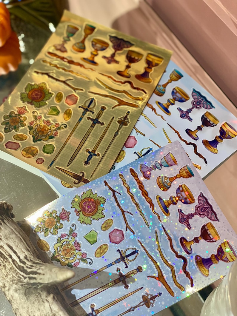 May include: Sheets of decorative stickers featuring tarot card suit symbols. The stickers include chalices, swords, wands, and pentacles in gold, blue, and iridescent finishes. The stickers are arranged on a light background.