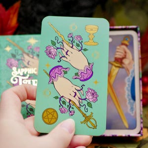 May include: A teal tarot card featuring two hands adorned with purple lace cuffs and holding a wand entwined with pink roses. The card also displays a golden chalice and a pentacle. The card is from the "Sapphic Tarot" deck.