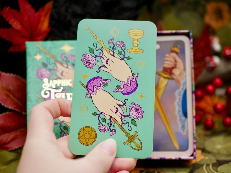 May include: A teal tarot card featuring two hands adorned with purple lace cuffs and holding a wand entwined with pink roses. The card also displays a golden chalice and a pentacle. The card is from the "Sapphic Tarot" deck.