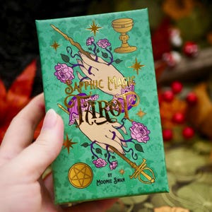 Puede incluir: Una caja de cartas del tarot verde con letras e ilustraciones doradas. La caja presenta las palabras "Sapphic Magic Tarot" e imágenes de manos, rosas, un cáliz y una varita. La caja está sostenida en una mano.