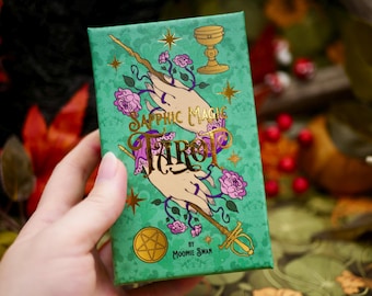 May include: A green tarot card box with gold lettering and illustrations. The box features the words "Sapphic Magic Tarot" and images of hands, roses, a chalice, and a wand. The box is held in a person's hand.