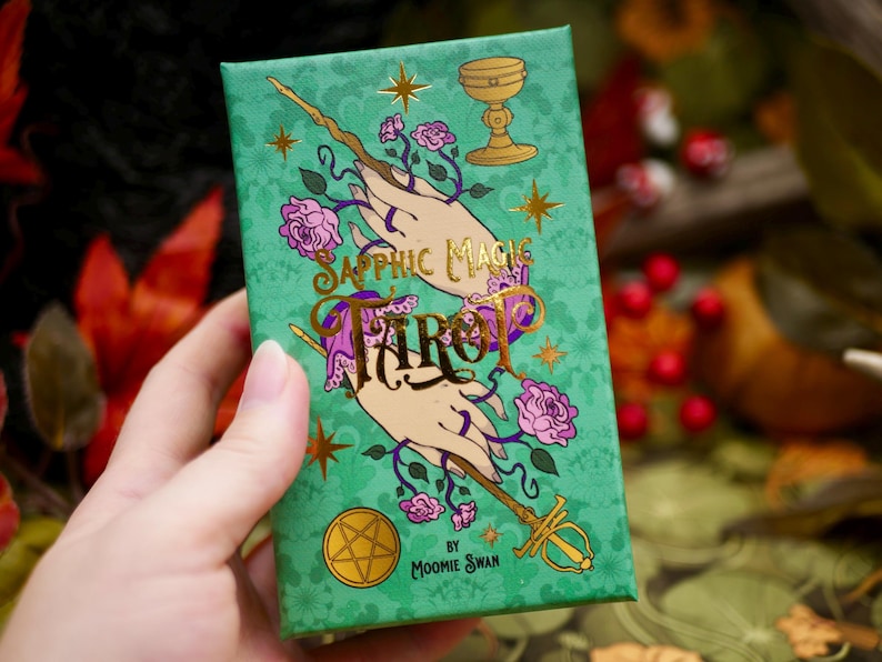 May include: A green tarot card box with gold lettering and illustrations. The box features the words "Sapphic Magic Tarot" and images of hands, roses, a chalice, and a wand. The box is held in a person's hand.