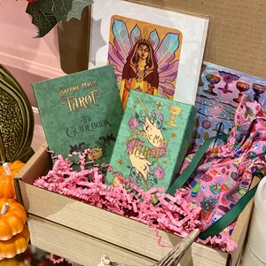 May include: A wooden box filled with a tarot card set, including a guidebook and deck, with pink paper filler. The set includes a print of a woman with a crown and a pink patterned bag. Decorative pumpkins and a candle are in the foreground.