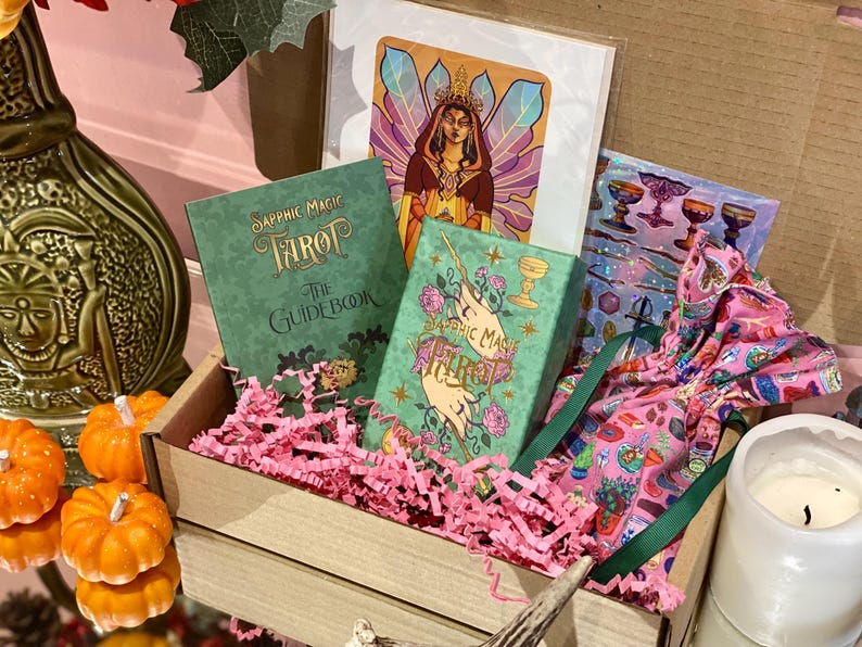 May include: A wooden box filled with a tarot card set, including a guidebook and deck, with pink paper filler. The set includes a print of a woman with a crown and a pink patterned bag. Decorative pumpkins and a candle are in the foreground.