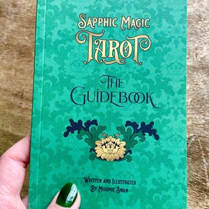 May include: A teal-colored tarot guidebook titled "Sapphic Magic Tarot: The Guidebook." The cover features ornate gold lettering and a floral design. The text "Written and Illustrated by Moomie Swan" is also visible.