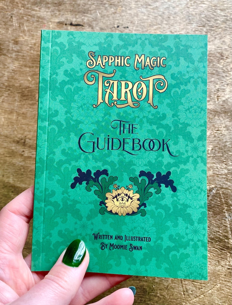 May include: A teal-colored tarot guidebook titled "Sapphic Magic Tarot: The Guidebook." The cover features ornate gold lettering and a floral design. The text "Written and Illustrated by Moomie Swan" is also visible.