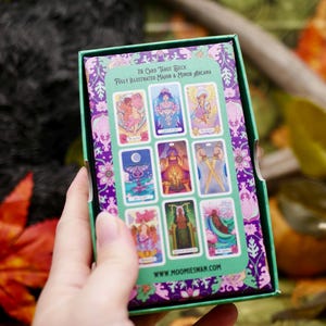 May include: A tarot card deck box with a green and purple floral design. The box displays a selection of illustrated tarot cards. The text on the box reads "78 Card Tarot Deck Fully Illustrated Major & Minor Arcana" and "www.moomieswan.com".