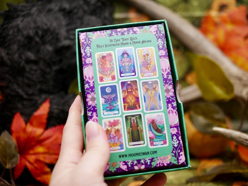 May include: A tarot card deck box with a green and purple floral design. The box displays a selection of illustrated tarot cards. The text on the box reads "78 Card Tarot Deck Fully Illustrated Major & Minor Arcana" and "www.moomieswan.com".