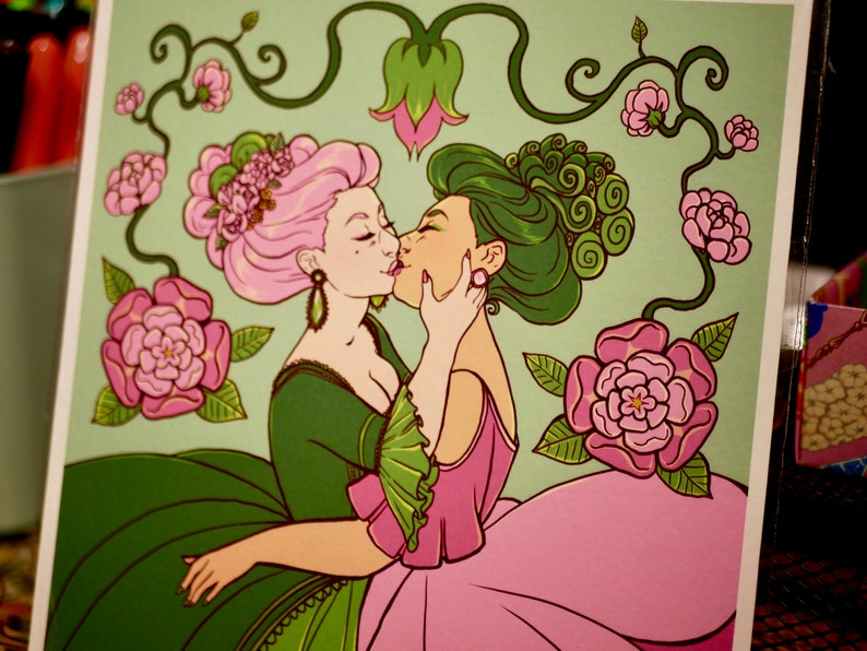 May include: Two women with pink and green hair are kissing. They are wearing elaborate dresses with floral patterns. The background is a light green with vines and pink flowers.