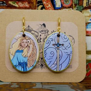 May include: Oval-shaped earrings with gold-colored hooks. One earring features a woman with an owl, the other a sword against a blue sky. The earrings are on a tan card with the text "SHOP SAPPHIRE DESIGNS".