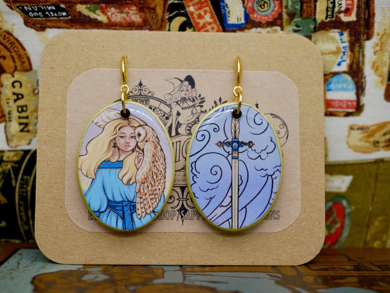 May include: Oval-shaped earrings with gold-colored hooks. One earring features a woman with an owl, the other a sword against a blue sky. The earrings are on a tan card with the text "SHOP SAPPHIRE DESIGNS".